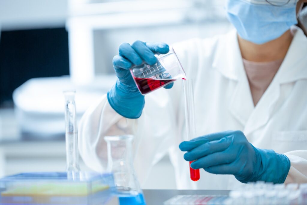 A scientist wearing rubber gloves and a white coat transports a translucent liquid between vials.